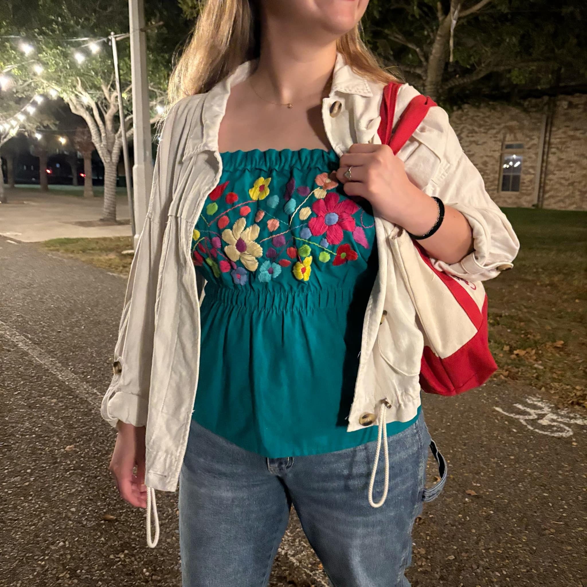 Image of May include: A teal embroidered peasant top with colorful flowers. The top is strapless and has a smocked waist. It is paired with a cream-colored utility jacket and blue jeans.