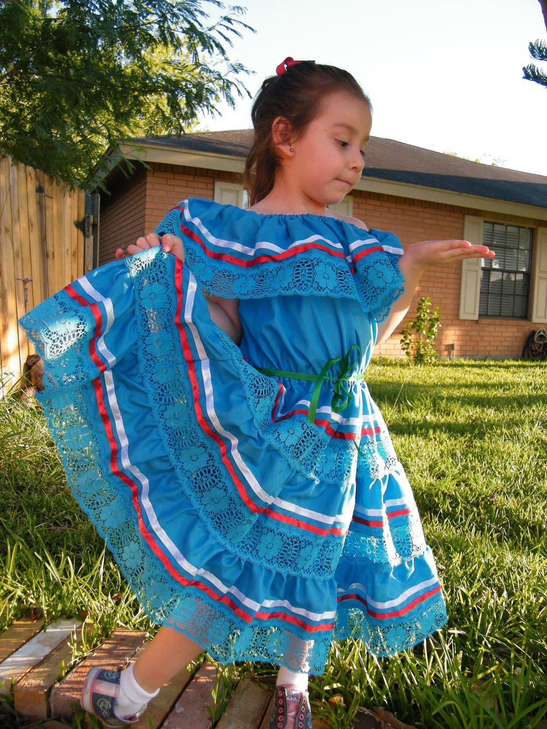 May include: A young girl wearing a blue and white dress with red and white ribbon trim. The dress has a lace overlay and is layered with ruffles. The girl is standing on a wooden surface with green grass in the background.
