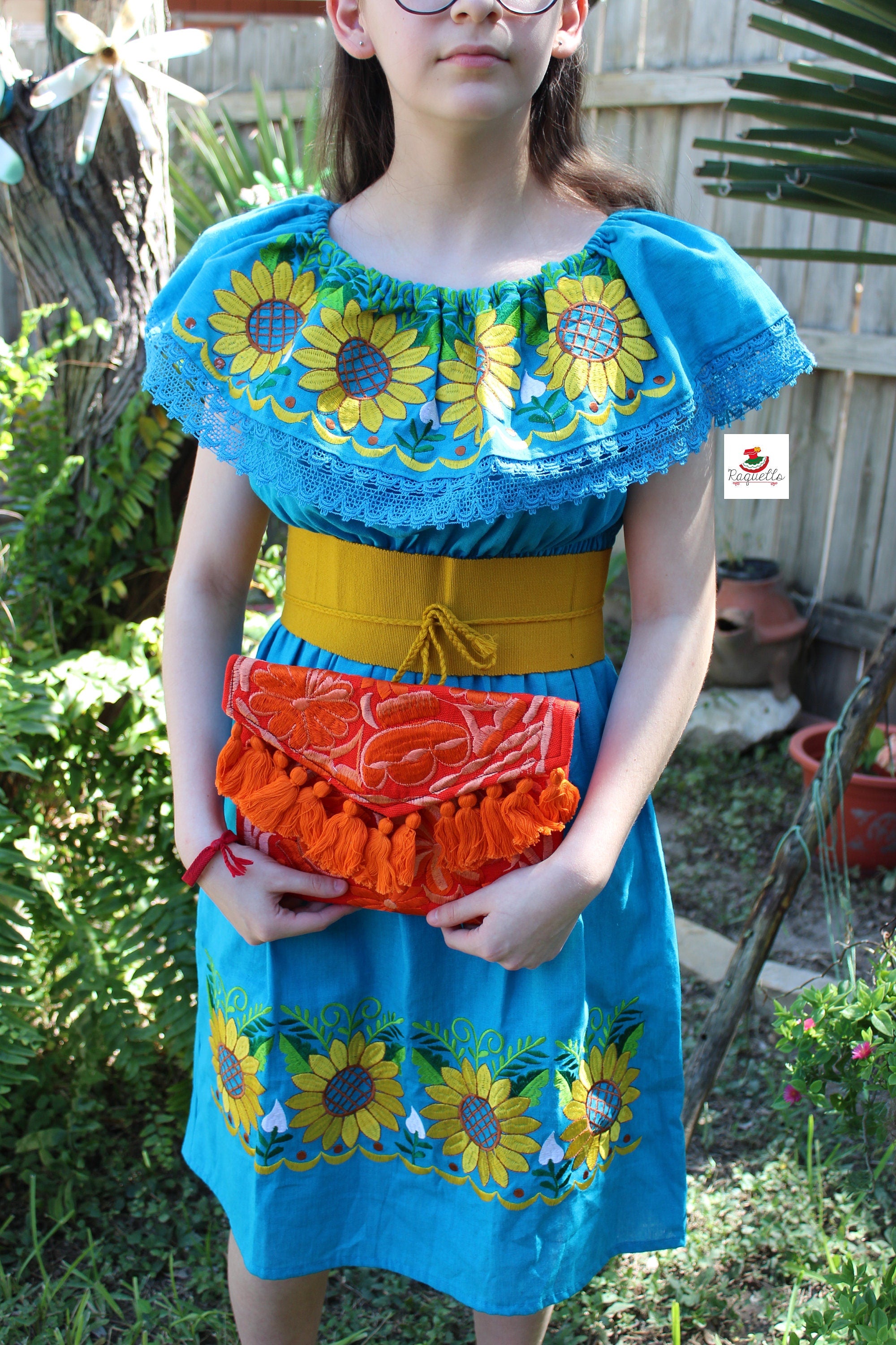 Image of May include: A blue embroidered dress with yellow sunflowers and a yellow belt. The dress has a ruffled neckline with white lace trim. The dress is paired with an orange embroidered clutch with orange tassels.