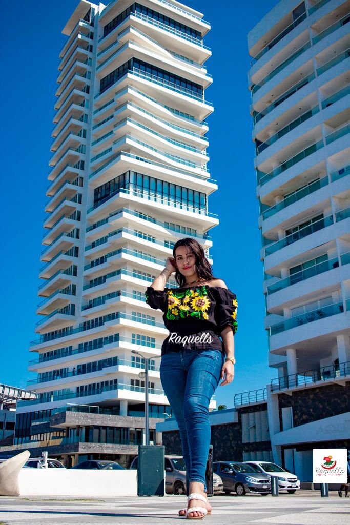 Image of May include: A woman wearing a black off-the-shoulder blouse with a sunflower embroidery design. The blouse has a black belt with the word 'Raquells' in white text. The woman is wearing blue jeans and white sandals. The woman is standing in front of two tall white buildings with balconies.